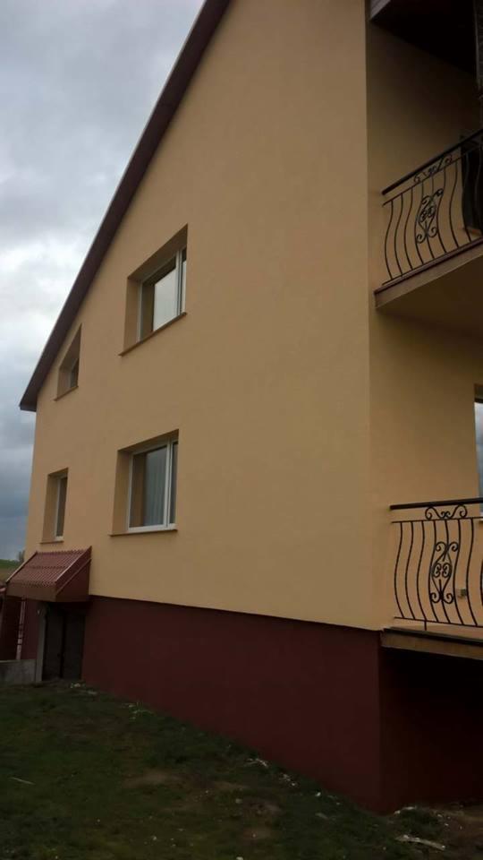 Dwupiętrowy dom z beżową elewacją, ciemnoczerwoną podmurówką i balkonami z kutymi balustradami, sfotografowany w pochmurny dzień.