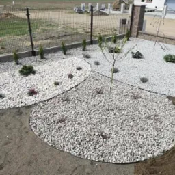 Nowo założony ogród z białym grysem, niskimi iglakami i młodymi drzewkami, widoczne obrzeża rabat i świeżo przygotowany grunt.