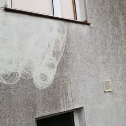 Szara elewacja budynku z widocznymi śladami po czyszczeniu, okna z białymi ramami i kratka wentylacyjna.