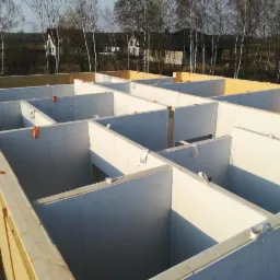 Wnętrze domu w trakcie budowy z widoczną konstrukcją ścian działowych i zewnętrznych, połączonych elementami z drewna i płyt OSB, z widocznymi mocowaniami i oznaczeniami na ścianach.