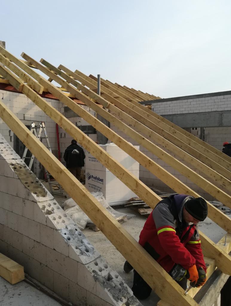 Montaż drewnianej konstrukcji dachu na ścianach z betonu komórkowego, widoczny pracownik w czerwonej kurtce używający wkrętarki, w tle materiały budowlane i drabina.