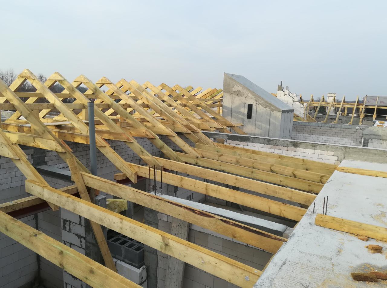 Konstrukcja drewniana dachu na budynku w trakcie budowy w Kołobrzegu, widoczne białe bloczki, elementy betonowe i wystające pręty zbrojeniowe.