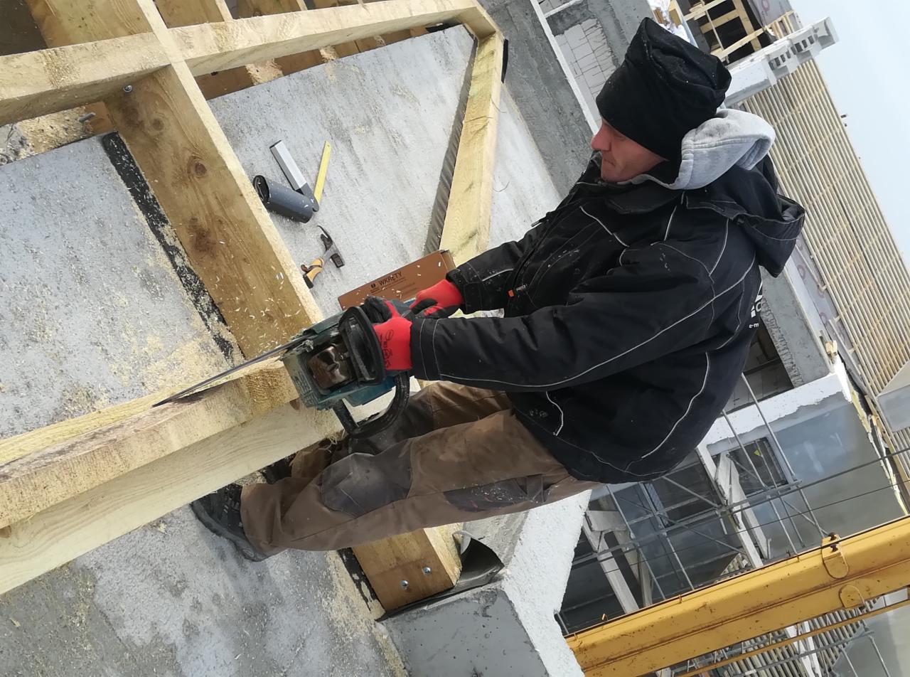 Pracownik w czapce i kurtce tnie drewniany element piłą łańcuchową na budowie domu, widoczne narzędzia i elementy konstrukcji.