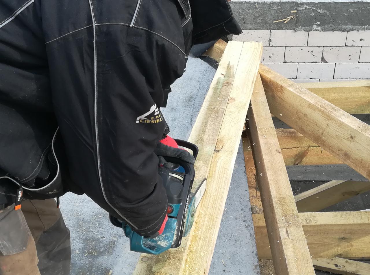 Pracownik w czarnej kurtce z logo firmy ciesielskiej tnie piłą łańcuchową drewniane elementy konstrukcji dachu na tle ściany z bloczków.