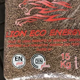 Worek 15 kg z brązowym pelletem drzewnym, z czerwonym logo lwa, certyfikatami ENplus i DINplus, leżący na czarnej nawierzchni z kostki brukowej.