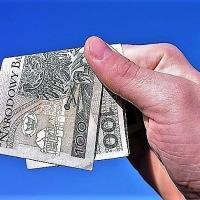 Dłoń trzymająca kilka zmiętych banknotów studolarowych na tle błękitnego nieba.