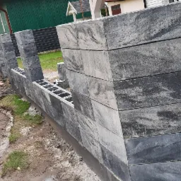 Wznoszenie ogrodzenia z szarych, betonowych bloczków z pustakami, widoczne słupki i fragmenty muru, na tle drogi i zaparkowanego samochodu.