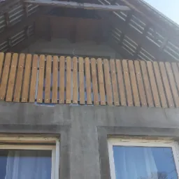 Surowy stan elewacji domu z drewnianą balustradą balkonu, widoczne okna z białymi ramami, brak wykończenia.