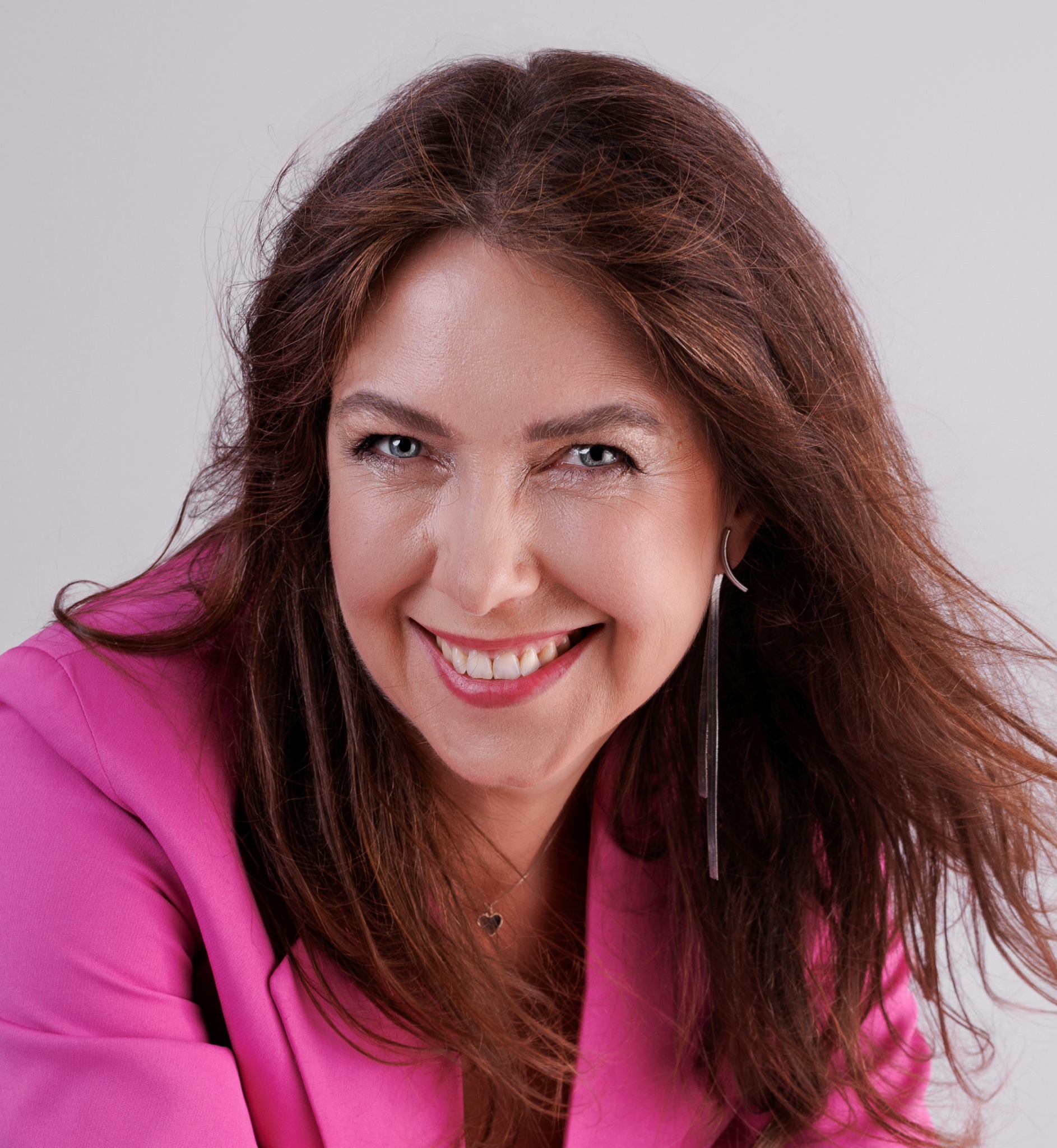 Uśmiechnięta kobieta w różowej marynarce, z długimi brązowymi włosami i srebrnymi kolczykami, patrzy w obiektyw na jasnym tle. Widoczny delikatny naszyjnik z serduszkiem.