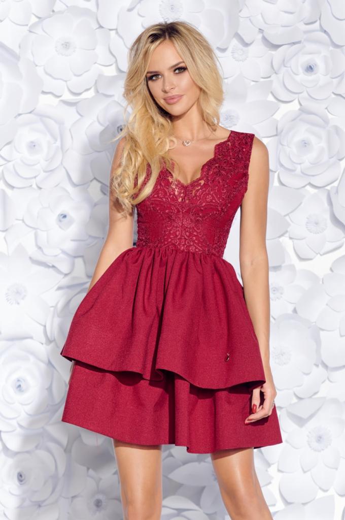 Kobieta w bordowej, dwuwarstwowej sukience z koronkową górą na tle białych, papierowych kwiatów.