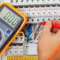 Pomiar napięcia na wyłącznikach różnicowoprądowych w rozdzielnicy elektrycznej za pomocą multimetru z pomarańczową obudową i czerwoną sondą pomiarową.