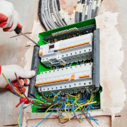 Montaż bezpieczników w skrzynce rozdzielczej, widoczne kable w kolorowej izolacji i ręce w roboczych rękawicach z czerwonymi elementami, dokręcanie śrubki w zabezpieczeniu.