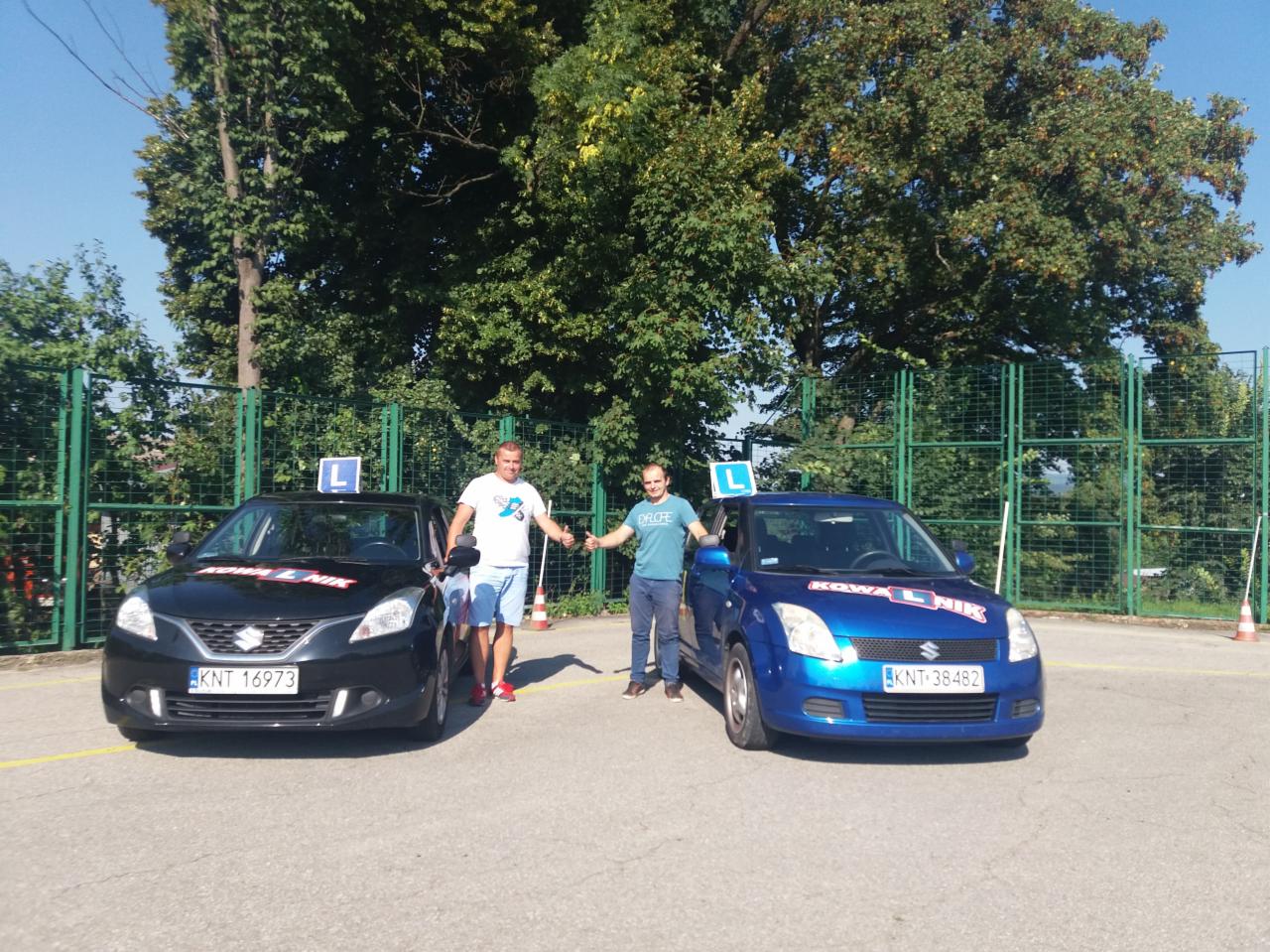 Instruktorzy pozują obok czarnego i niebieskiego samochodu z literą L na dachu, na placu manewrowym, w tle zielone drzewa i ogrodzenie.