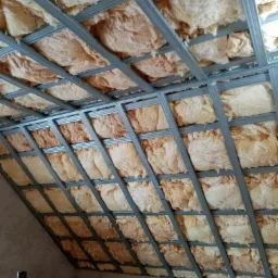 Skośny sufit poddasza z widoczną konstrukcją metalową i wypełnieniem z wełny mineralnej, częściowo oświetlony, z rolką izolacji Isover w dolnym rogu.