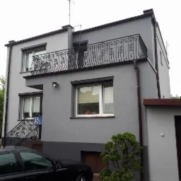 Dwukondygnacyjny dom z szarą elewacją, balkonem z ozdobną balustradą i brązowymi rynnami, widoczny numer domu 39A, częściowo zasłonięty przez czarny samochód.