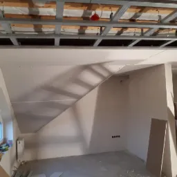 Montaż płyt kartonowo-gipsowych na skośnym poddaszu, widoczne metalowe profile sufitowe, fragment okna dachowego i przygotowane płyty gipsowe oparte o ścianę.