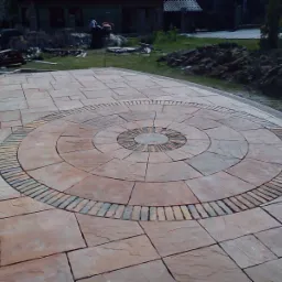 Wykończony taras z kostki brukowej w odcieniach brązu, z centralnym elementem dekoracyjnym w kształcie koła, otoczony regularną kostką, w tle widoczny trawnik i budynek.