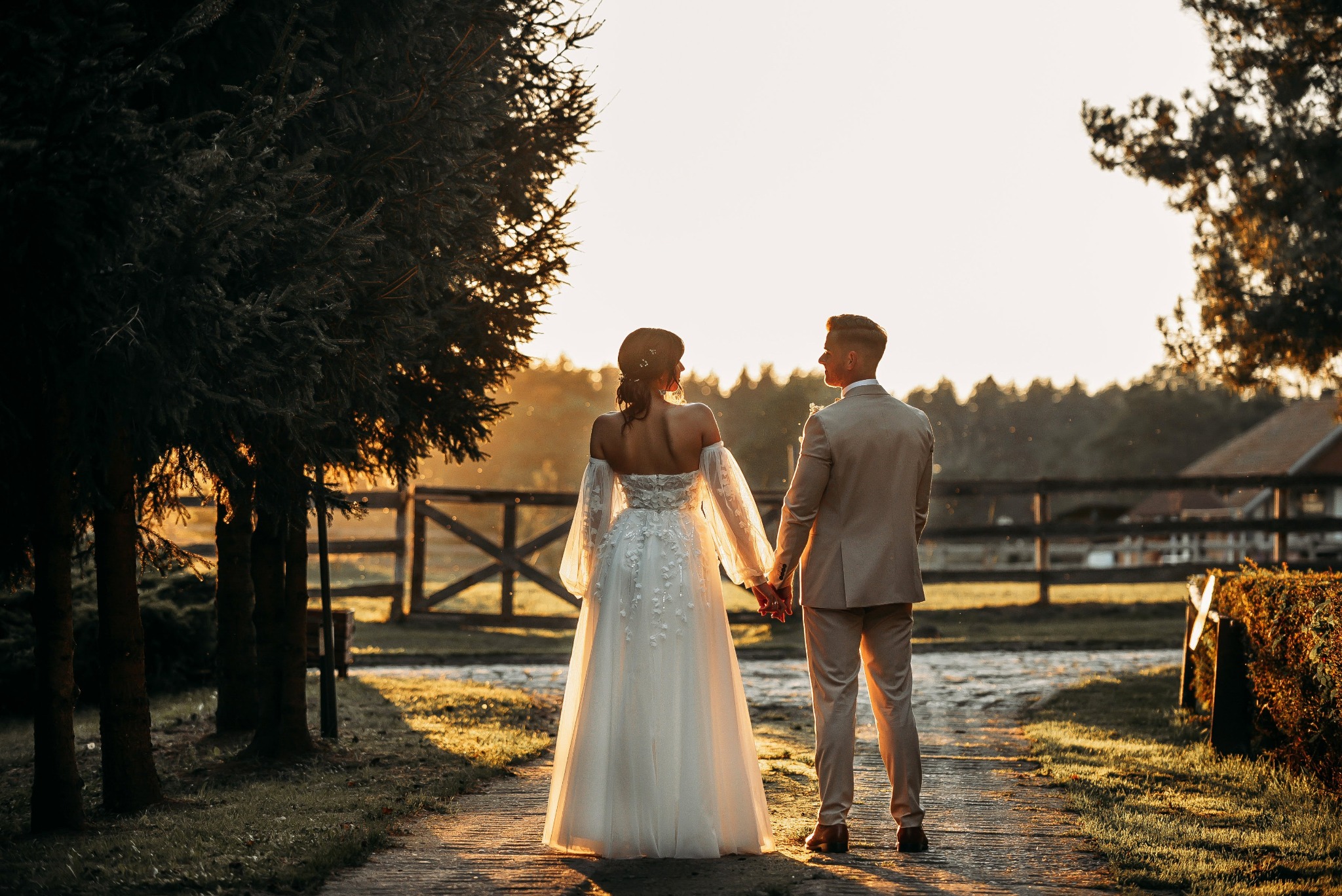 Para młoda, trzymając się za ręce, idzie ścieżką w kierunku zachodzącego słońca, widok z tyłu, ona w sukni z odkrytymi ramionami i długimi, przezroczystymi rękawami, on w beżowym garniturze.