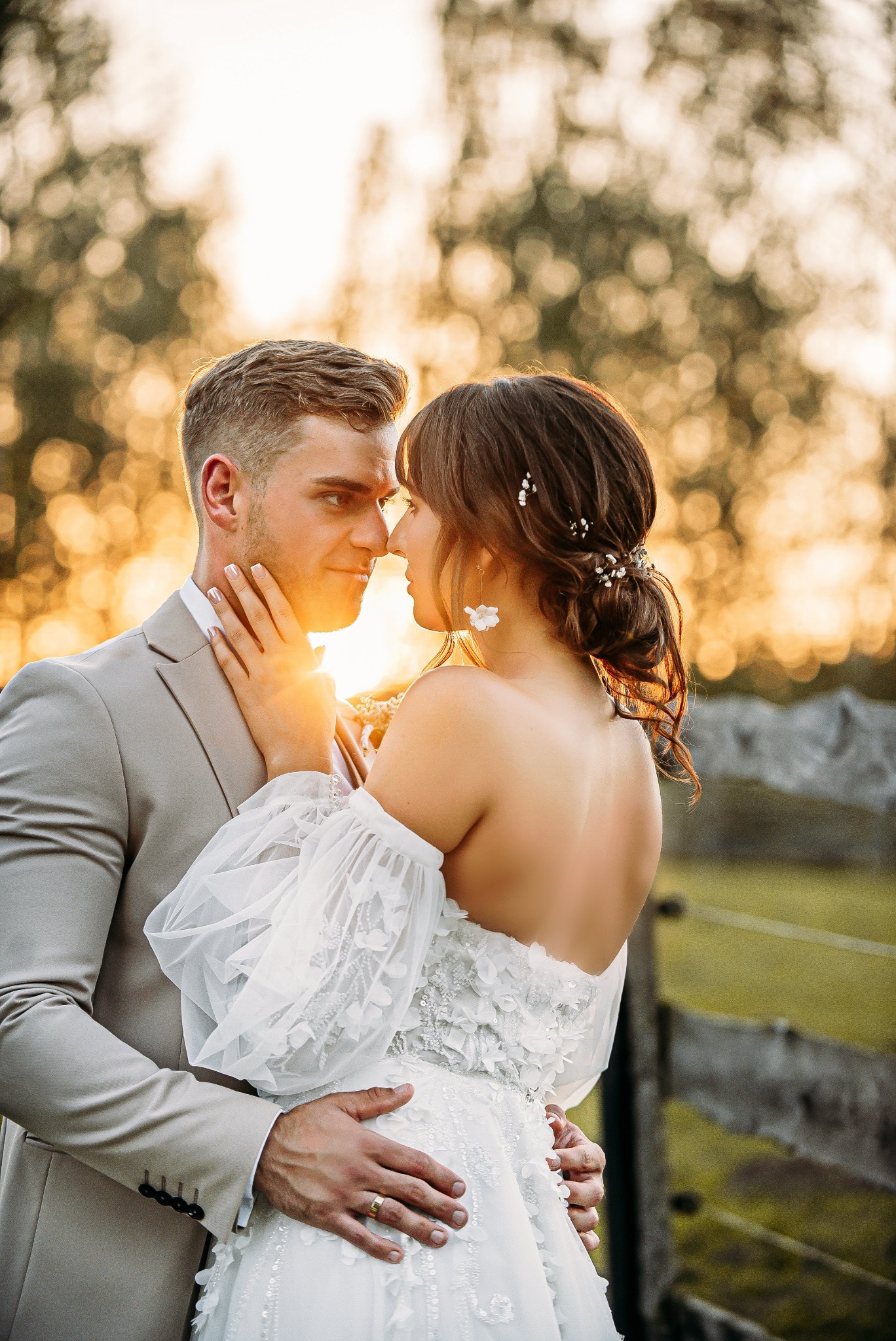 Para młoda w objęciach, ona w sukni z aplikacjami kwiatowymi i bufkami, on w beżowym garniturze, na tle rozmytego, złotego światła zachodzącego słońca.