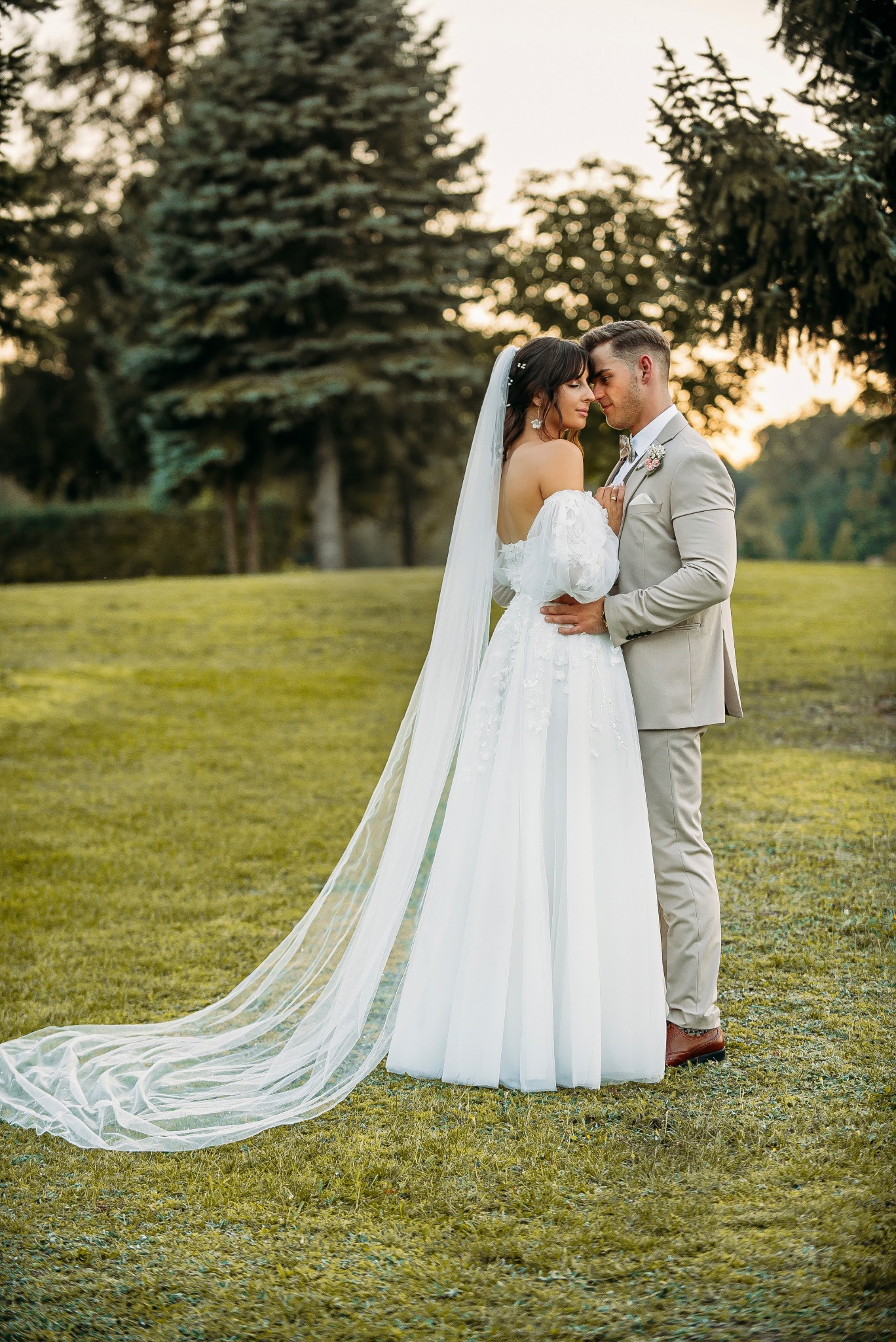Para młoda w objęciach na zielonym trawniku, panna młoda w białej sukni z długim welonem, pan młody w beżowym garniturze, drzewa w tle.