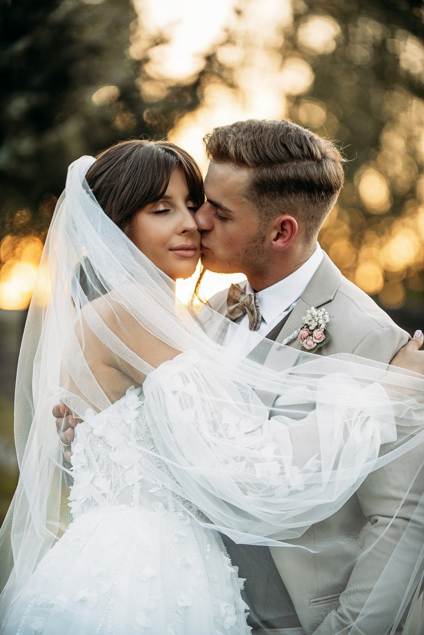 Panna młoda w koronkowej sukni i welonie, z zamkniętymi oczami, jest całowana w policzek przez pana młodego w beżowym garniturze z butonierką, na tle rozmytego, ciepłego światła zachodzącego słońca.