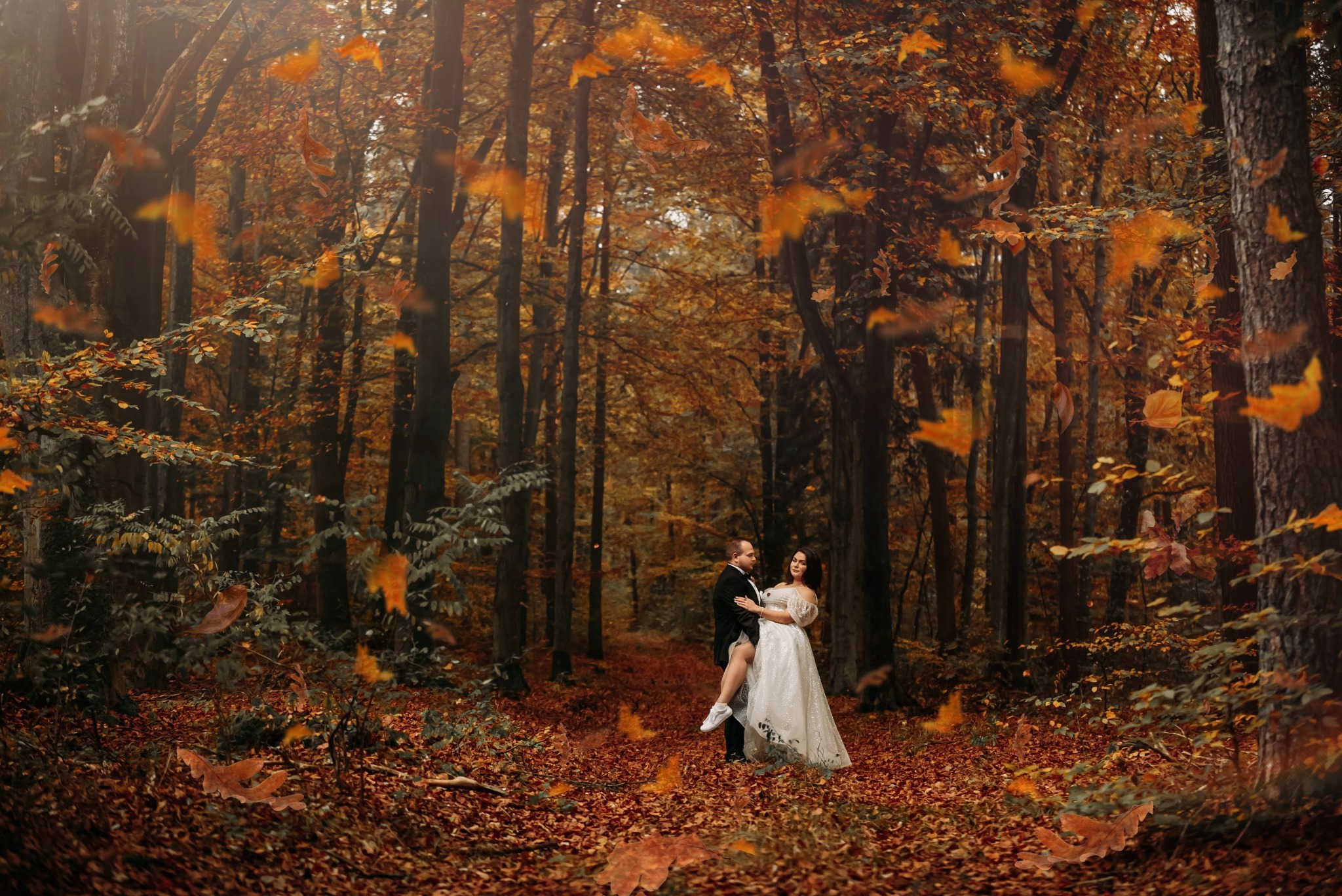 Para młoda w lesie jesienią; mężczyzna w czarnym smokingu trzyma pannę młodą w białej sukni, unosząc ją lekko w górę, podczas gdy opadają liście.