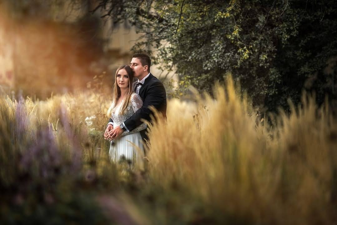 Para młoda w objęciach na tle pola trawy ozdobnej, pan młody w czarnym smokingu całuje pannę młodą w czoło, ona w białej sukni z koronkowymi rękawami.