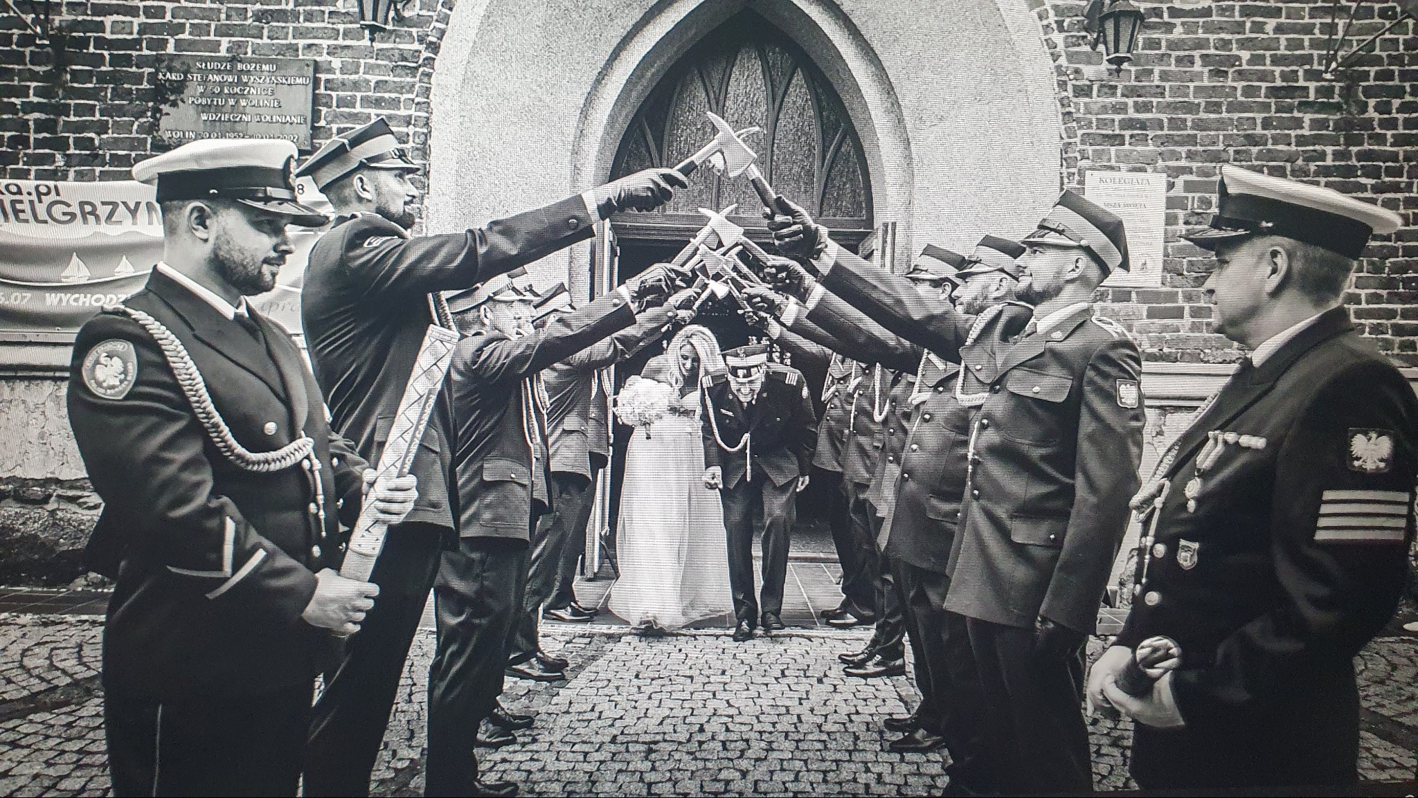 Czarno-białe zdjęcie nowożeńców przechodzących pod szpalerem strażaków w mundurach trzymających toporki nad głowami, przed ceglanym budynkiem z tablicą pamiątkową.