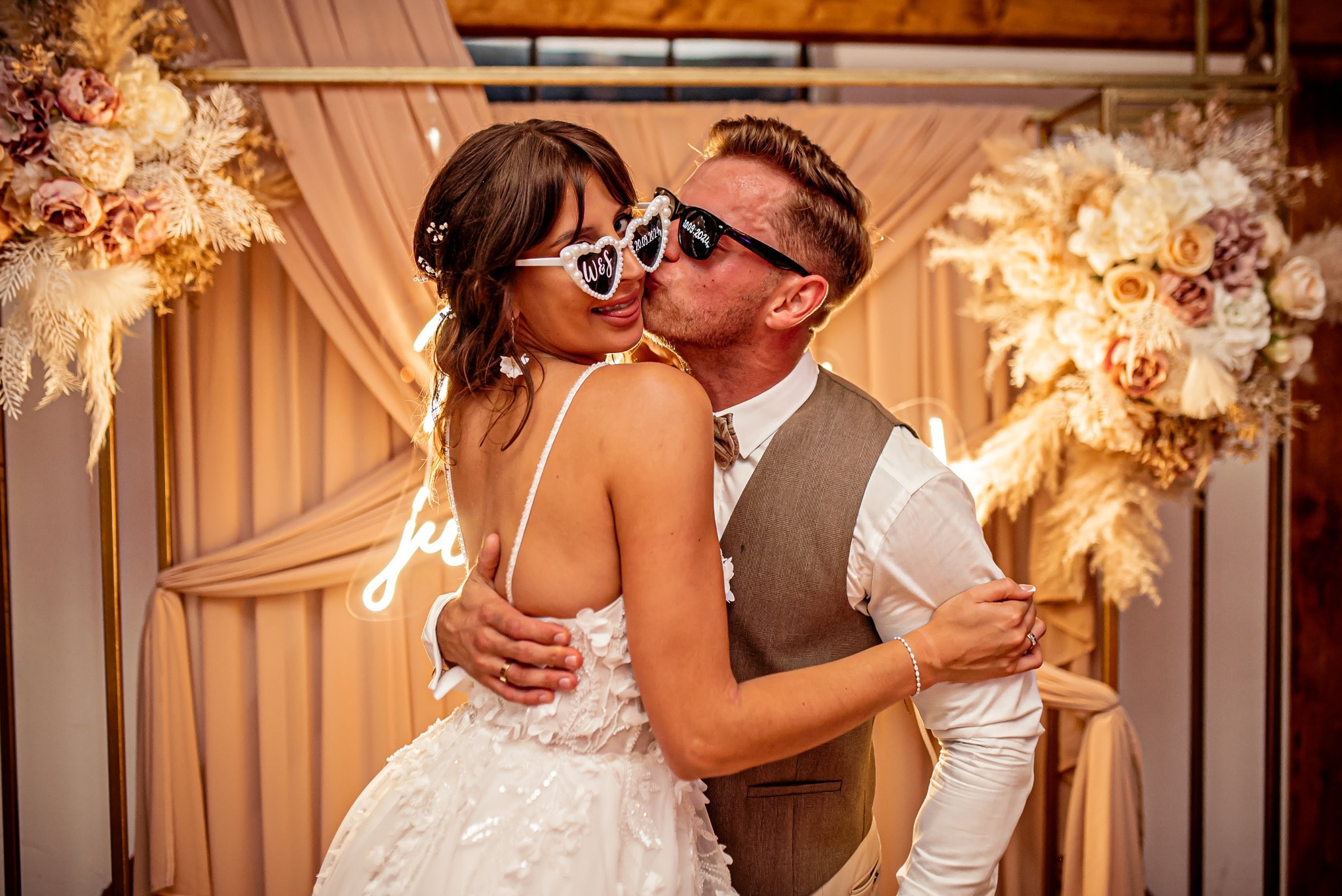 Panna młoda w białej sukni i okularach w kształcie serc z napisem 'W&S 20.08.2024' całowana w policzek przez pana młodego w okularach z datą '16.08.2024' na tle dekoracji ślubnej z kwiatów...