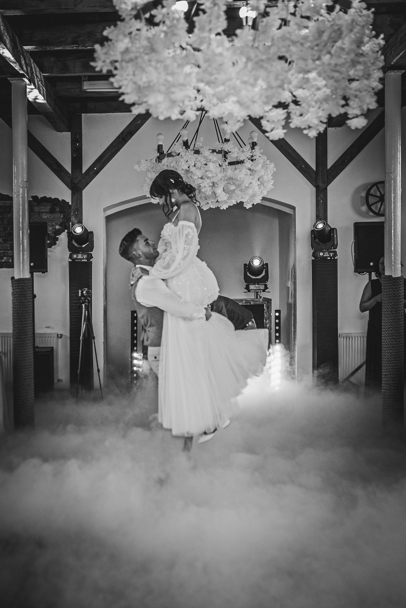 Czarno-białe zdjęcie pary młodej podczas pierwszego tańca, unoszącej się w dymie pod dekoracyjnym żyrandolem z kwiatów, z widocznymi reflektorami i kamerą na statywie w tle.