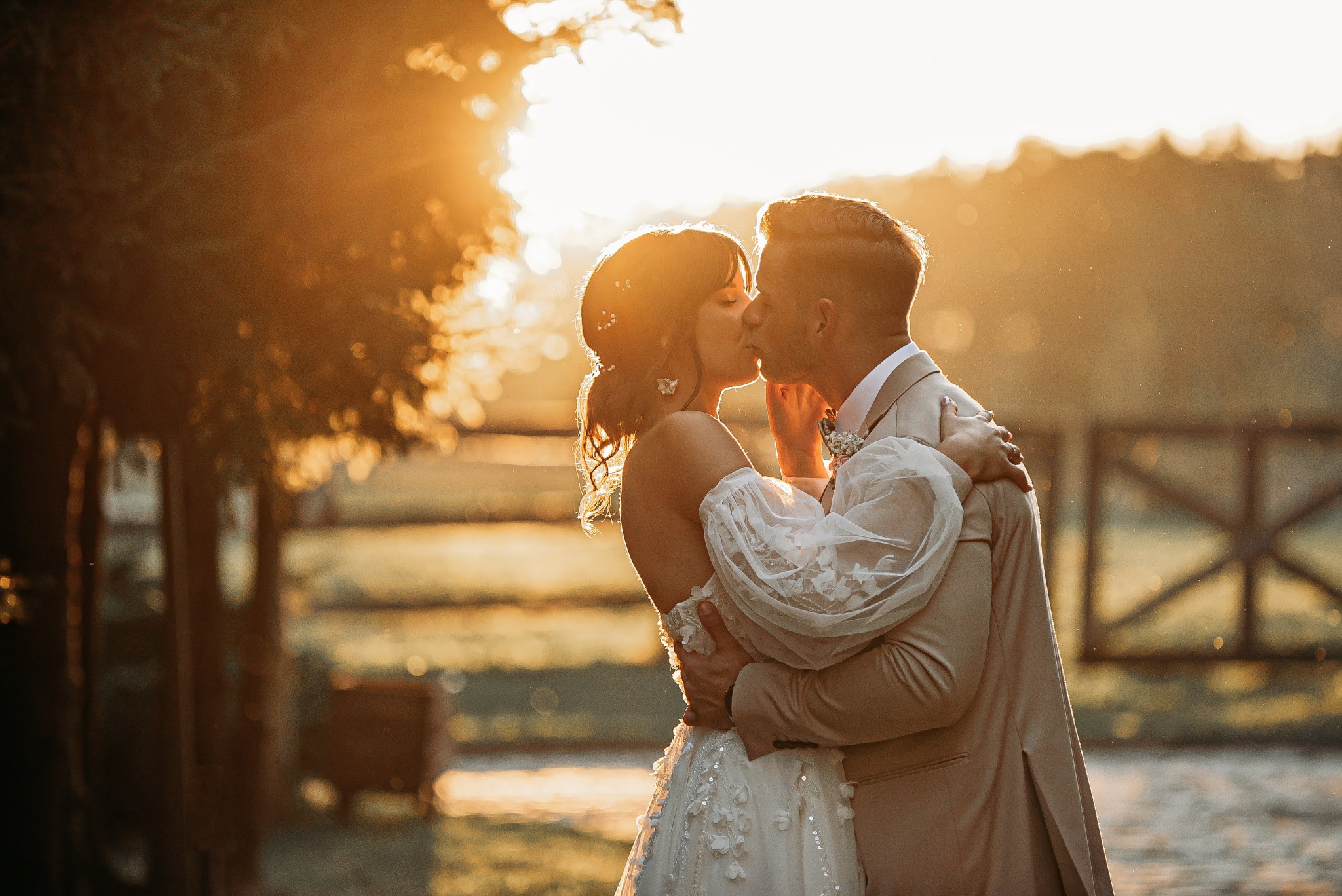 Para młoda w objęciach, pocałunek na tle zachodzącego słońca, ona w sukni z haftowanymi kwiatami i bufkami, on w beżowym garniturze, drewniane ogrodzenie w tle.