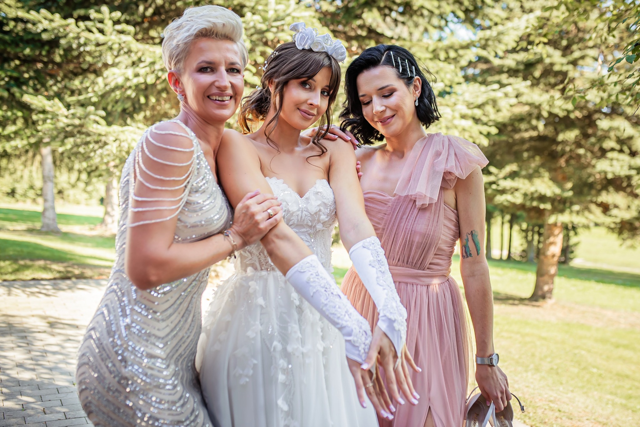 Panna młoda w białej sukni ślubnej z kwiatowym wiankiem, pozująca z dwiema druhnami w sukniach wieczorowych na tle zieleni parkowej.