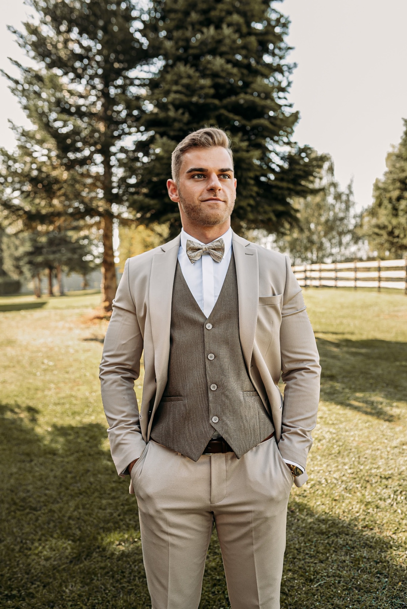 Elegancki mężczyzna w beżowym garniturze i muszce, pozujący na tle zielonego trawnika i drzew podczas słonecznego dnia.