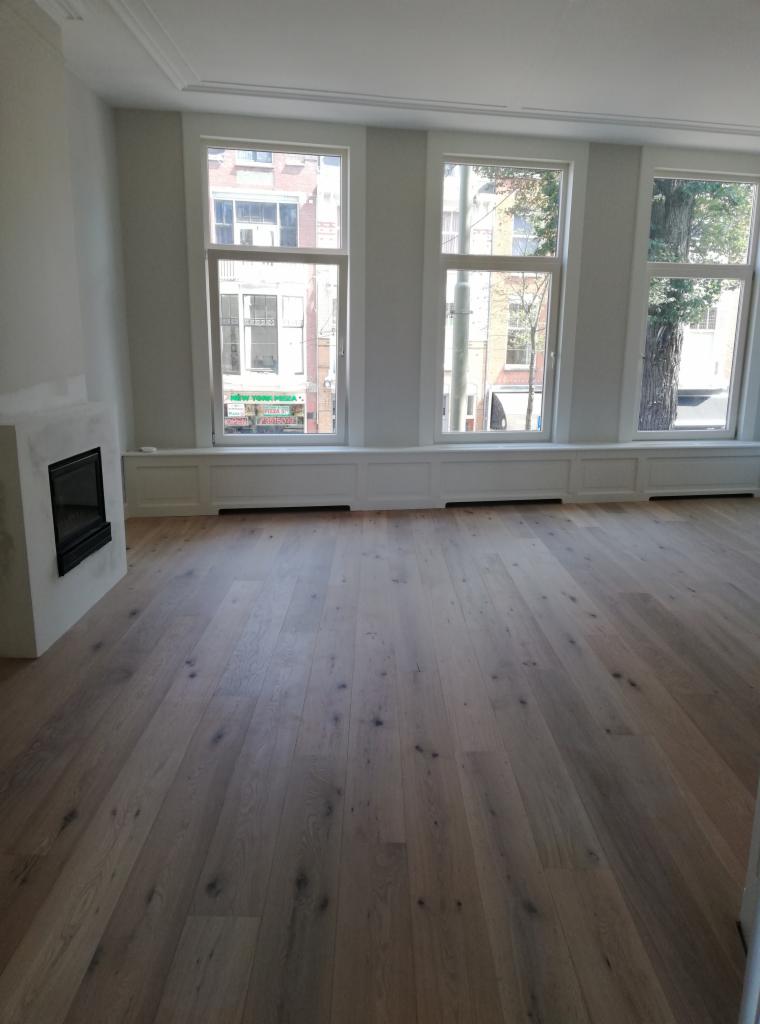 Jasne panele podłogowe w przestronnym pokoju z trzema oknami i kominkiem z surowego betonu, widok na ulicę z budynkami.