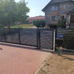 Nowoczesna, pozioma, czarna brama przesuwna z metalowymi panelami, widoczna skrzynka pocztowa i tabliczka ostrzegawcza 'Uwaga zły pies', na tle domu z cegły i kostki brukowej.