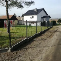 Zielona siatka panelowa ogrodzenia posesji z podmurówką betonową, widok z polnej drogi na dom z ciemnym dachem i bramką do piłki nożnej w ogrodzie.