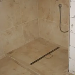 Beżowa kabina prysznicowa z odpływem liniowym, wykończona płytkami imitującymi kamień, widoczne elementy instalacji wodnej i fragment surowej ściany.