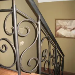 Kuta, metalowa balustrada schodowa z dekoracyjnymi zawijasami, zamontowana na brązowych, ceramicznych schodach. Na ścianie wisi obraz.