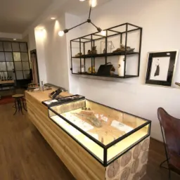 Wnętrze sklepu z drewnianą ladą ekspozycyjną z przeszkloną gablotą, oświetloną od spodu, oraz metalową półką na ścianie, oświetloną lampą z żarówkami Edison