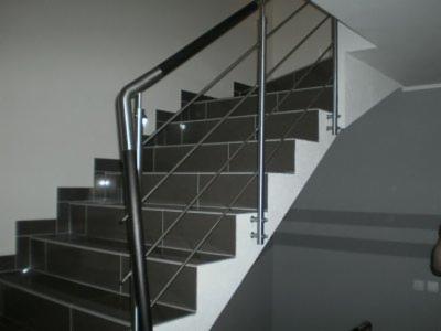 Schody wyłożone ciemnymi płytkami z balustradą ze stali nierdzewnej, z czarną poręczą, w nowoczesnym wnętrzu domu. Ujęcie z dołu, ukazujące detale wykończenia.