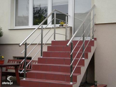 Zewnętrzne schody z czerwoną terakotą i nowoczesną balustradą ze stali nierdzewnej, prowadzące do drzwi wejściowych. Widoczny fragment okna i ławki.