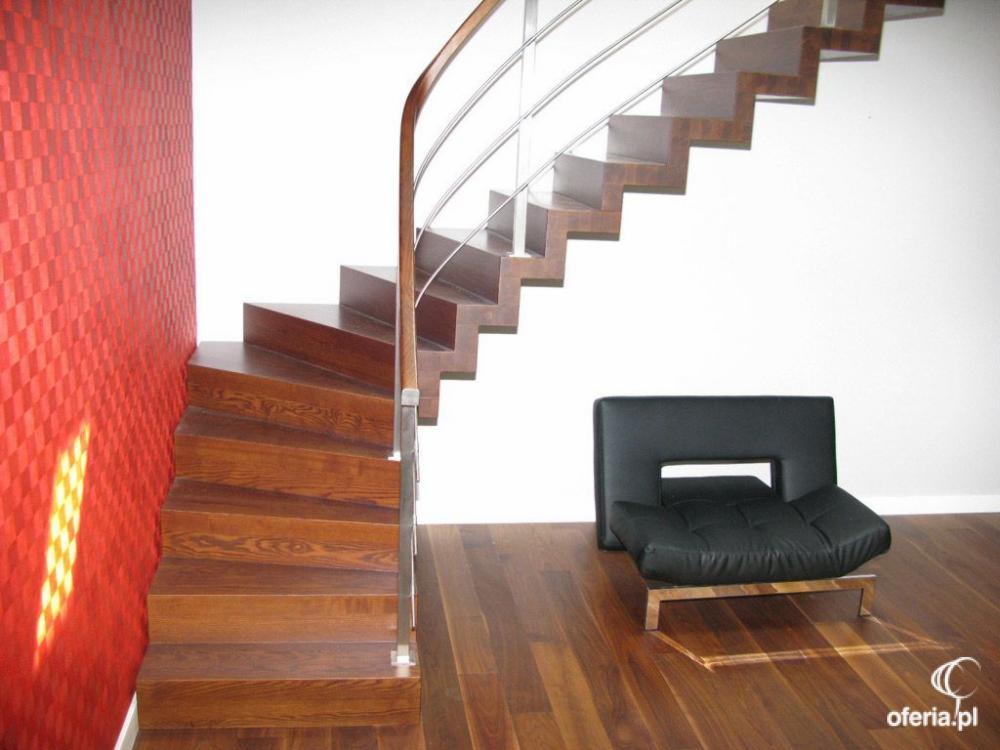 Drewniane schody z metalową balustradą i poręczą, prowadzące do góry. Czerwona ściana z geometrycznym wzorem. Na podłodze leży czarny fotel.