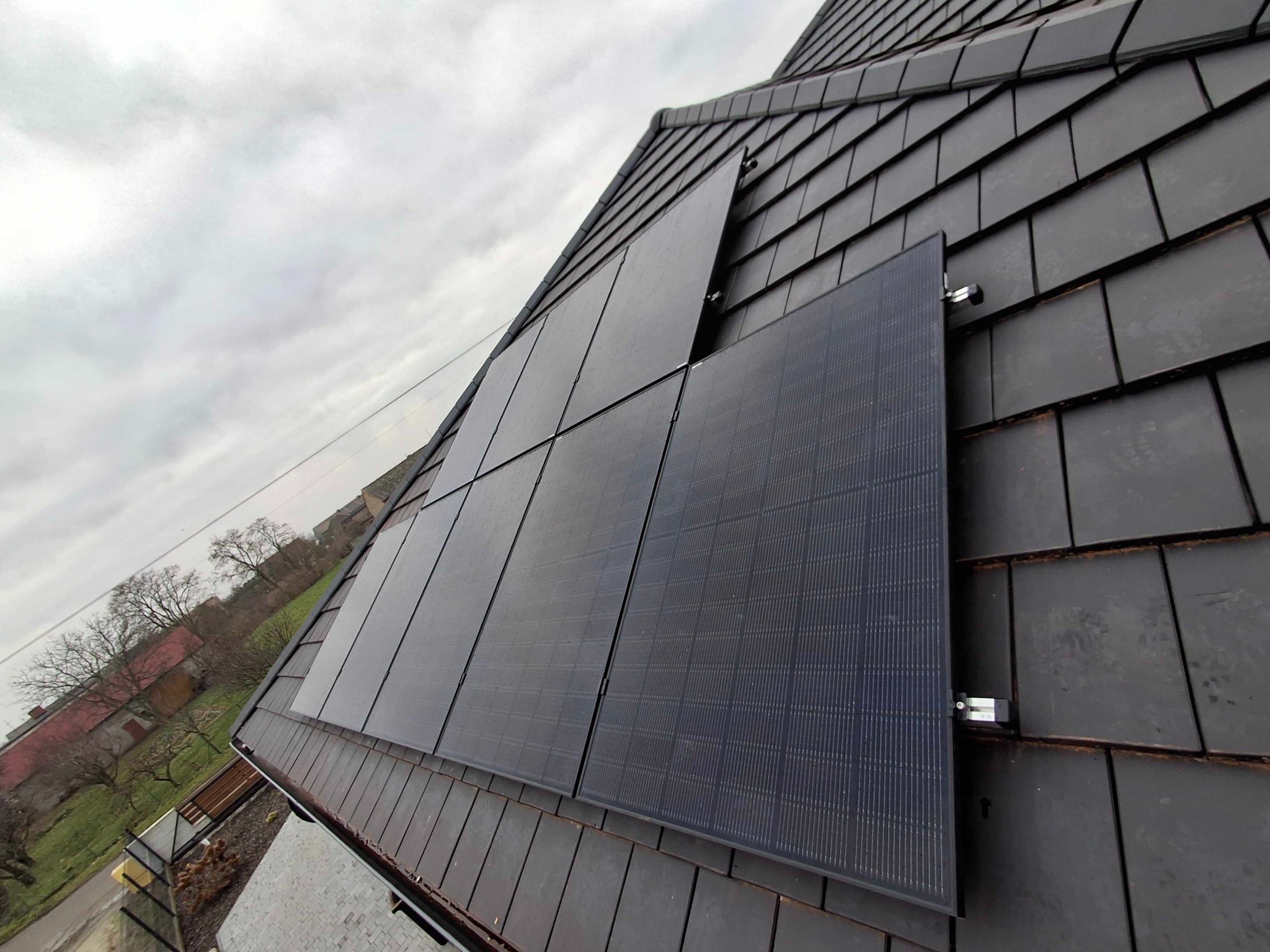 Panele słoneczne zamontowane na dachu pokrytym ciemną dachówką, widok z dołu pod kątem, pochmurne niebo w tle.