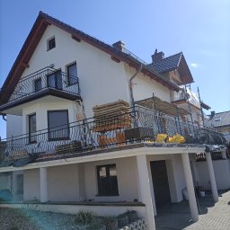 Wiesław Motyl - Dwupiętrowy dom z balkonami w trakcie prac elewacyjnych. Widoczne rusztowania, materiały budowlane i świeżo pomalowana fasada. Słoneczny dzień, błękitne niebo.