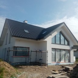Wiesław Motyl - Nowoczesny dom jednorodzinny z ciemną dachówką i jasną elewacją w trakcie wykończenia. Widoczne rusztowania i zabezpieczone okna folią.