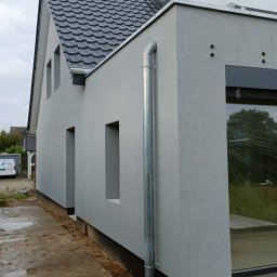 Wiesław Motyl - Szara elewacja domu jednorodzinnego z widoczną rynną spustową i fragmentem dachu z czarną dachówką. Nowoczesny styl, surowy krajobraz wokół budynku.