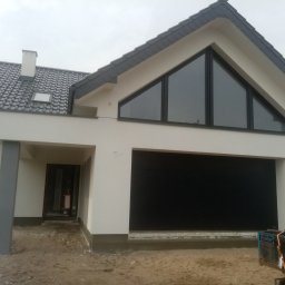 Wiesław Motyl - Nowoczesny dom jednorodzinny z garażem i dużym trójkątnym oknem na poddaszu. Biała elewacja, grafitowa dachówka i brama garażowa. Teren wokół domu w trakcie prac wykończeniowych.