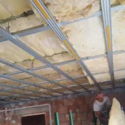 Montaż izolacji z wełny mineralnej na poddaszu, widoczne metalowe profile sufitowe, fragment ściany z czerwonej cegły i pracownik w tle.