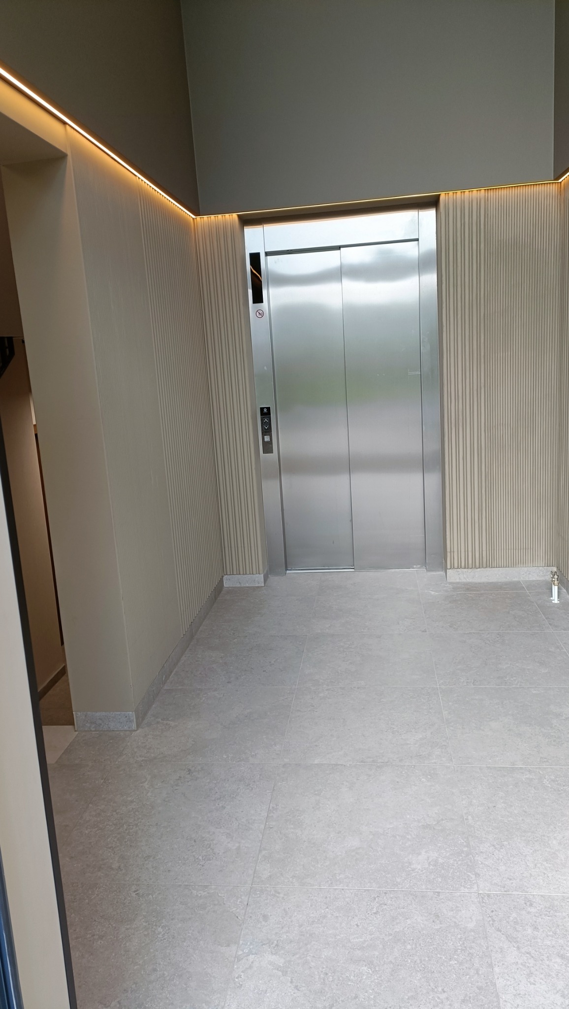 Nowoczesny hol z windą: ściany w beżowe pionowe pasy, podświetlenie LED u góry, szare płytki podłogowe. Minimalistyczny design, metalowe drzwi windy.