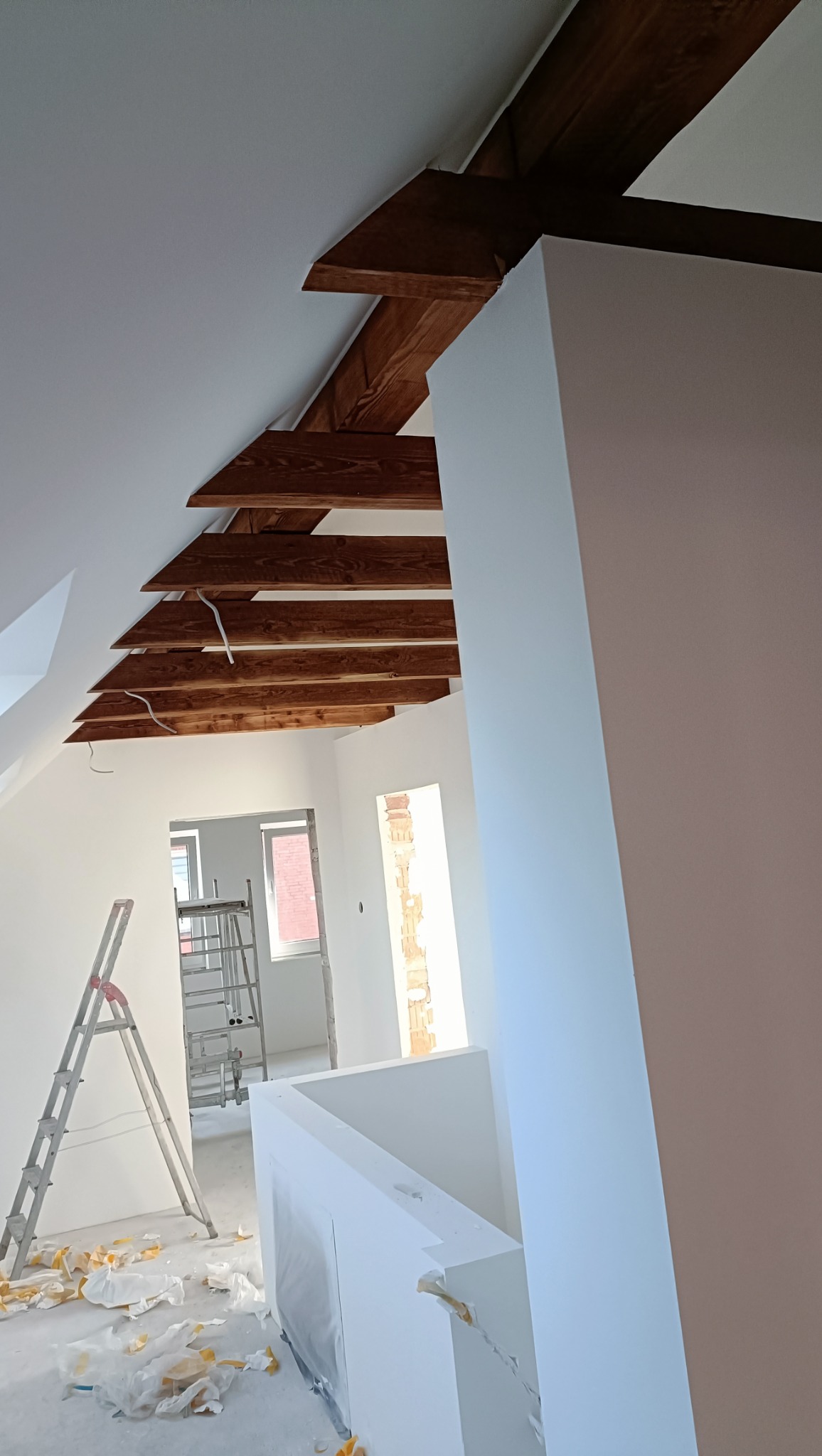 Białe ściany poddasza z drewnianymi belkami stropowymi, widoczne drabiny i resztki folii malarskiej na podłodze. W tle okno w stanie surowym.